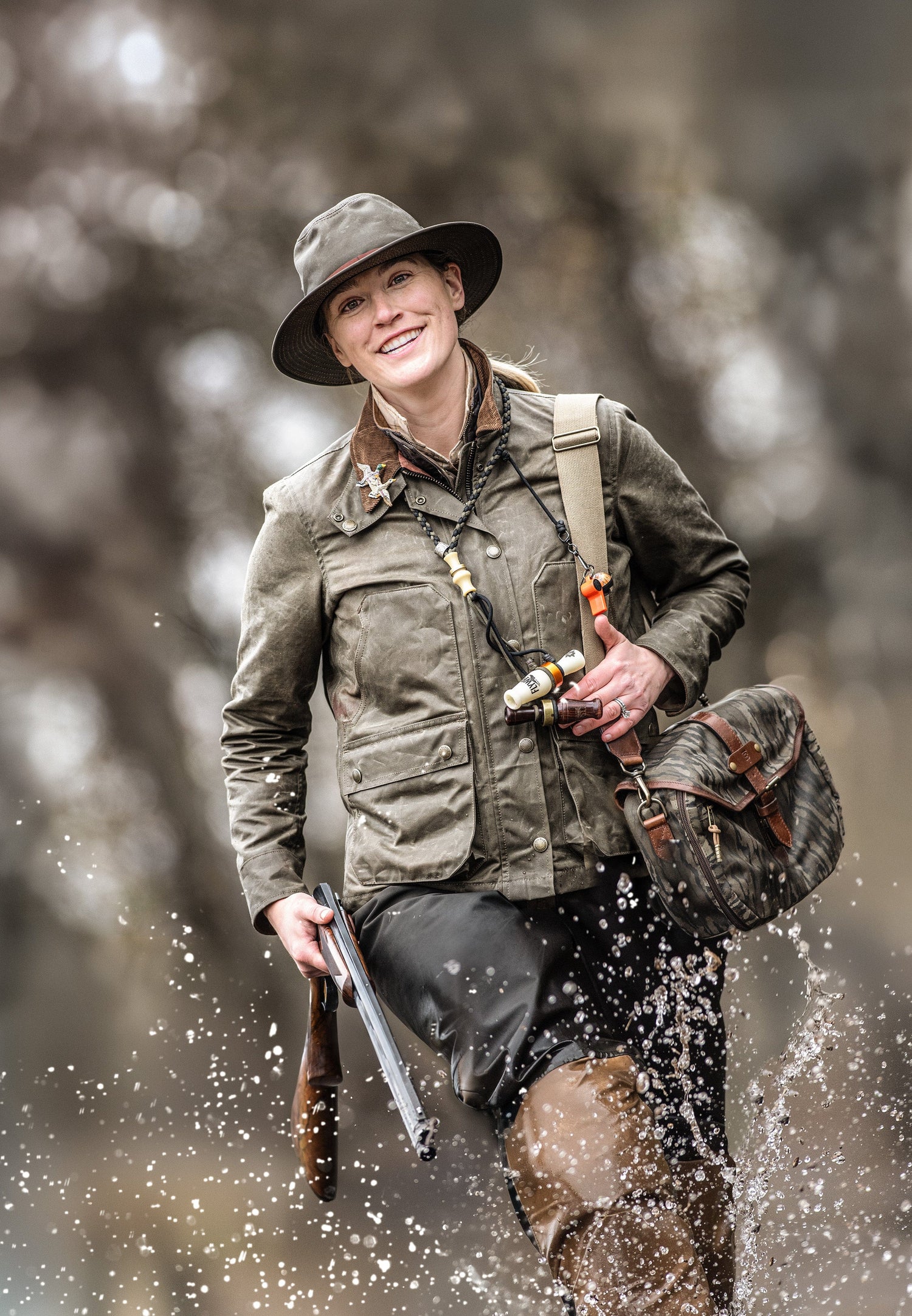 A person in outdoor gear and waders walks through water, holding a fishing rod and a shotgun, with a shoulder bag.