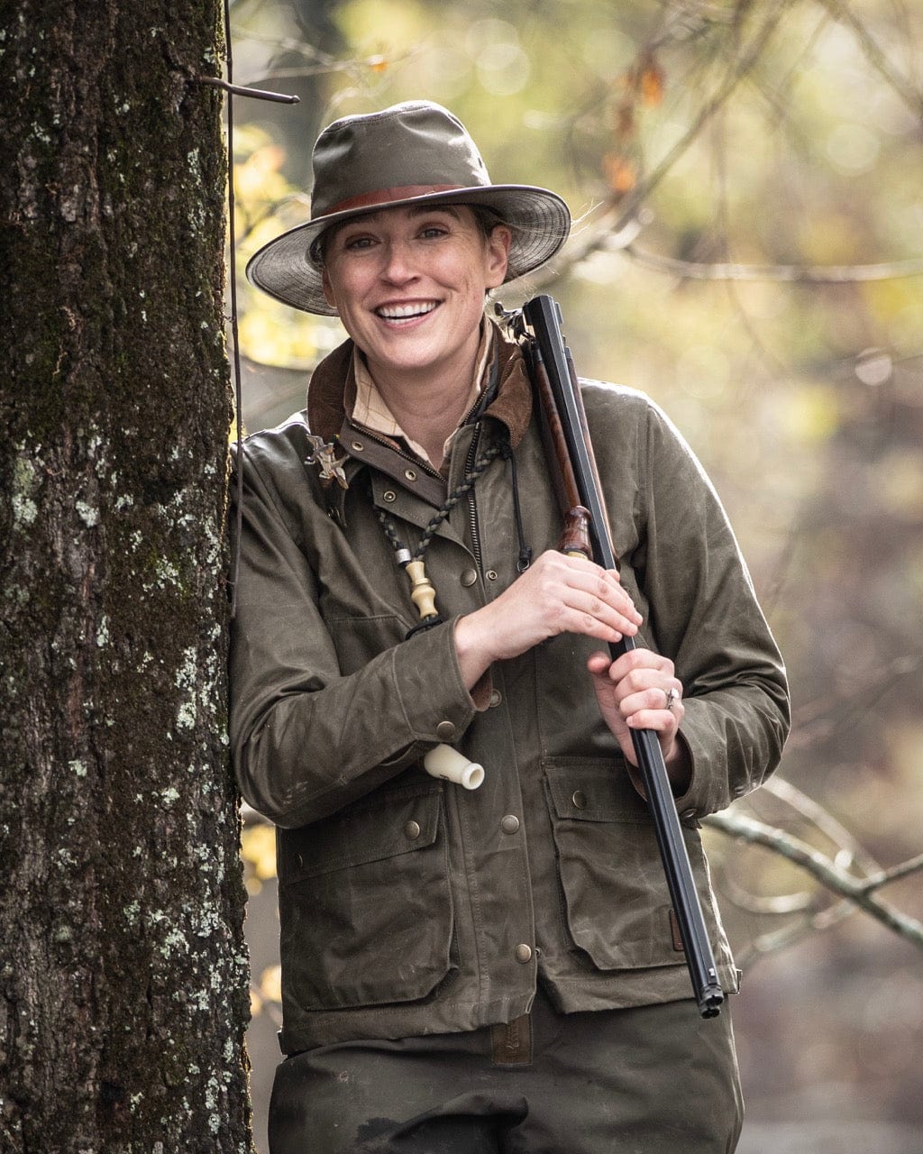 A person in outdoor clothing and hat stands by a tree, smiling and holding a shotgun in both hands.