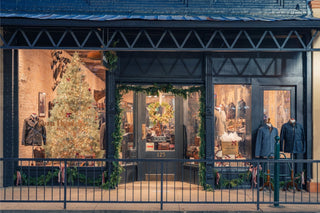 Clothing store window display with Christmas decorations, including a lit tree and greenery, featuring mens apparel and accessories.
