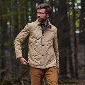 Man stands in the woods wearing a light tan quilted jacket with collar and hickory redmont pants.