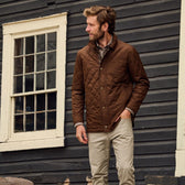 Man stands outside of a house wearing a brown quilted jacket with collar and khaki redmont pants.