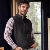Bearded man in a plaid shirt and Tom Beckbes Warren Fleece Vest, standing indoors by a window with trees outside.