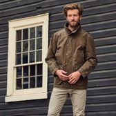 A man stands before a wooden building in a Tom Beckbe Tensaw Jacket and beige pants.