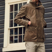 A person in a Tom Beckbe Tensaw Jacket stands with hands in pockets by a dark wooden building with a window.
