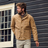 A man in a Tom Beckbe Fowler Jacket and light pants stands before a dark wooden building, looking to his left.
