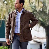 A man in a Tom Beckbe Piedmont Jacket strolls outdoors, hand in pocket, with trees and a building behind him.