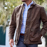 A person in a Tom Beckbe Piedmont Jacket over a blue checkered shirt stands outdoors in a leafy setting.