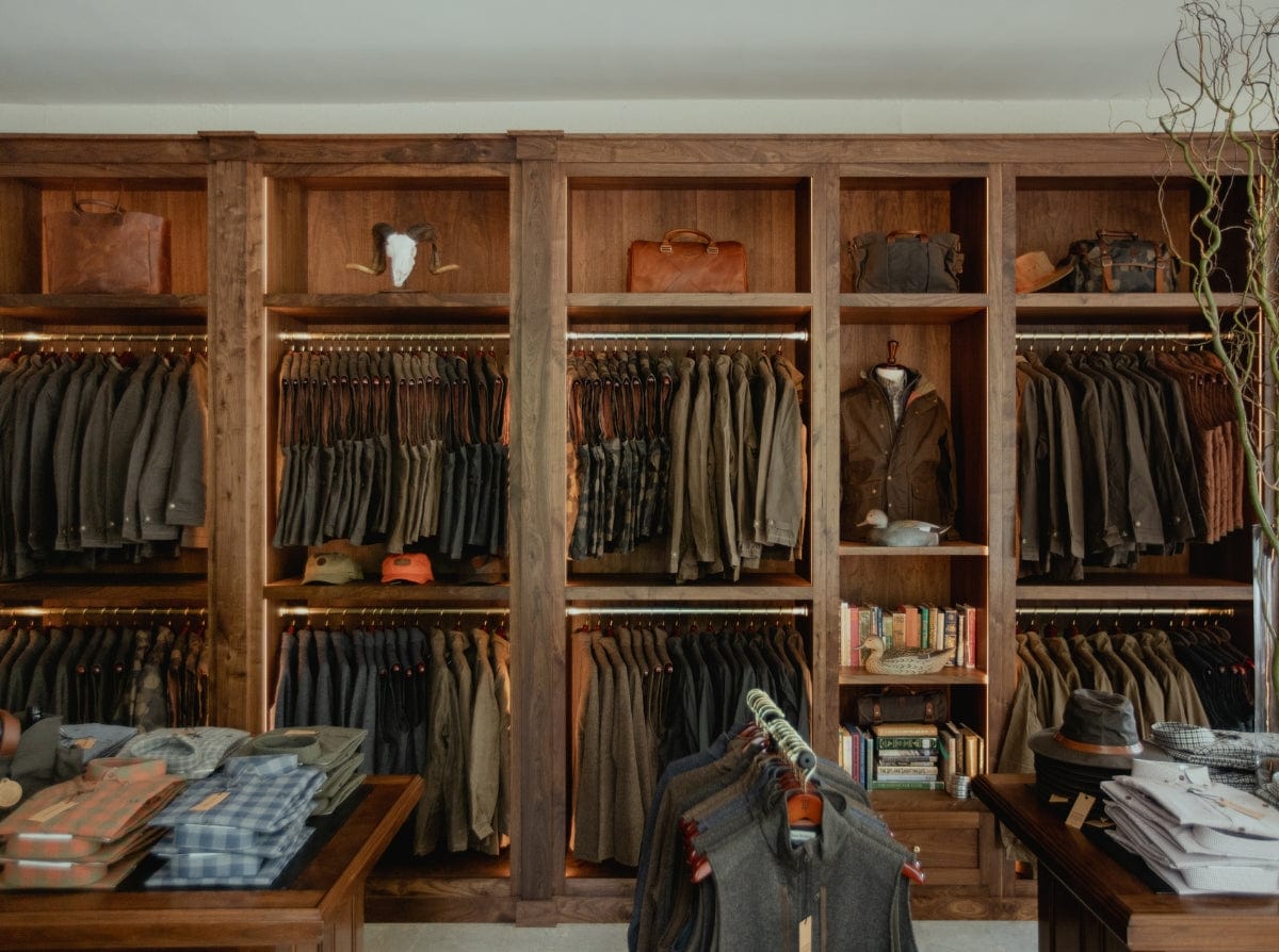 Clothing store interior with wooden shelves displaying assorted jackets and bags, and tables with folded shirts and hats.