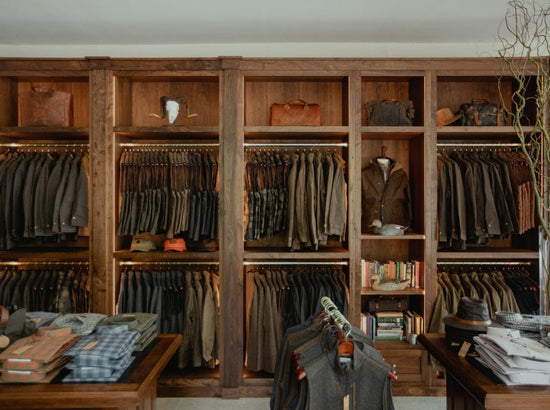Clothing store interior with wooden shelves displaying assorted jackets and bags, and tables with folded shirts and hats.