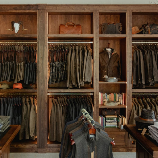 Clothing store interior with wooden shelves displaying jackets, hats, and books. Racks of shirts in the foreground.