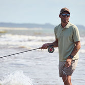 Man in a Tom Beckbe Coastal Polo Shirt, cap, and sunglasses fishing with a rod in shallow ocean waters with waves behind.