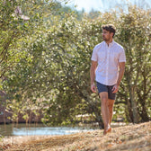 A man in a Tom Beckbe Tidewater Shirt and shorts strolls barefoot on grass near water, with trees in the background.