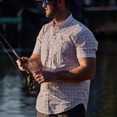 A person in a Tom Beckbe Tidewater Shirt and sunglasses is fly fishing by water, holding a fishing rod.