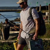 Man in a Tidewater Shirt by Tom Beckbe holds gear and tackle box at the waterfront, enjoying UPF 30+ coverage.
