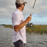 A person in a cap and sunglasses stands by water holding a fishing rod, wearing a Tom Beckbe Tidewater Short Sleeve Shirt.