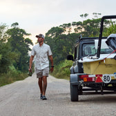 A man in a Tom Beckbe Tidewater Shirt (Short Sleeve) with UPF 30+ walks a dirt road by a parked car towing a boat.