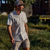 Man in a Tom Beckbe Tidewater Shirt and shorts stands outdoors, wearing a cap, enjoying the sun.