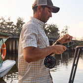 Man in a Tom Beckbe Tidewater Shirt and cap adjusts rod line by a river, boat and bridge in the background.