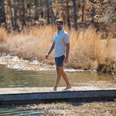A man in a Tom Beckbe Tidewater Shirt and shorts walks barefoot on a wooden dock by a lake, surrounded by tall grasses.