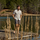 A man in a Tom Beckbe Tidewater Short Sleeve Shirt and shorts walks barefoot on a wooden dock by a lakeside with trees.