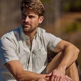 A man in a Tidewater short sleeve by Tom Beckbe sits outdoors in shorts, gazing into the distance with a neutral look.