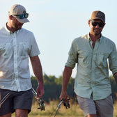 Two men in Tom Beckbe Tidewater Shirts and sunglasses walk outdoors, each holding fishing rods.