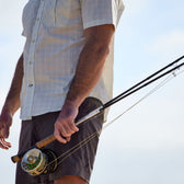 Person holding a fishing rod, wearing Tom Beckbes Tidewater Shirt, standing against a clear sky.
