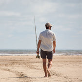 A man in a cap and shorts walks barefoot on a sandy beach, wearing a Tom Beckbe Tidewater Shirt, holding a fishing rod.