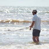 Man fishing in knee-deep ocean waves, wearing a Tom Beckbe Tidewater Shirt and cap.