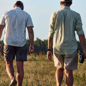Two men in Tom Beckbe Tidewater Shirts stroll through a grassy field with fishing rods, sunlight overhead, and trees behind them.