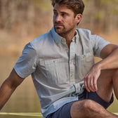 A man in a Tom Beckbe Tidewater Shirt (Short Sleeve) and shorts sits by the water, looking sideways.