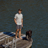 Barefoot man in a Tom Beckbe Tidewater Shirt (Short Sleeve), UPF 30+, stands on a dock with his black dog, water glimmering.