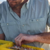 A person in a Tom Beckbe Tidewater Shirt inspects fishing flies in a yellow tackle box.
