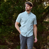 Man in a Tom Beckbe Tidewater Shirt (Short Sleeve), green plaid, and gray pants walking outdoors with trees behind.