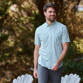 A person wearing a Tom Beckbe Tidewater Shirt and gray pants smiles near white wooden chairs outside.