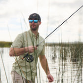 A man in a Tidewater Shirt by Tom Beckbe and sunglasses holds a fishing rod among tall reeds near water.