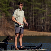 A man in a Tom Beckbe Tidewater Shirt fishes on a boat as his dog sits nearby, framed by the forested shoreline.