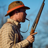 Man outdoors in plaid shirt, wearing Tom Beckbe Field Hat Band, holding shotgun, blowing whistle under clear blue sky.