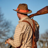 Man in a plaid shirt and Tom Beckbe Field Hat Band holds a rifle and bird against a clear blue sky.