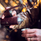 Man pours bourbon from a stainless bottle flask wrapped in leather into a stainless toasting cup.
