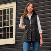 A woman in a Tom Beckbe Womens Fairmont Vest over a white shirt and jeans stands by a wooden building with a window.
