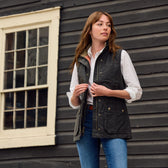 A woman in a white shirt and the Tom Beckbe Womens Fairmont Vest stands outside a dark wooden building, looking aside.