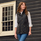A woman wearing a Tom Beckbe Womens Fairmont Vest and jeans stands before a dark wooden building with windows.