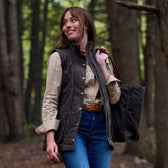 Woman in a plaid shirt and vest, carrying a bag in a forest, glances to the side with a smile.