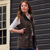 A woman in a Tom Beckbe Womens Fairmont Vest and white shirt stands thoughtfully before a dark wooden building.