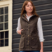 A woman in a Tom Beckbe Womens Fairmont Vest and white shirt stands by a dark wooden wall, looking to the side.